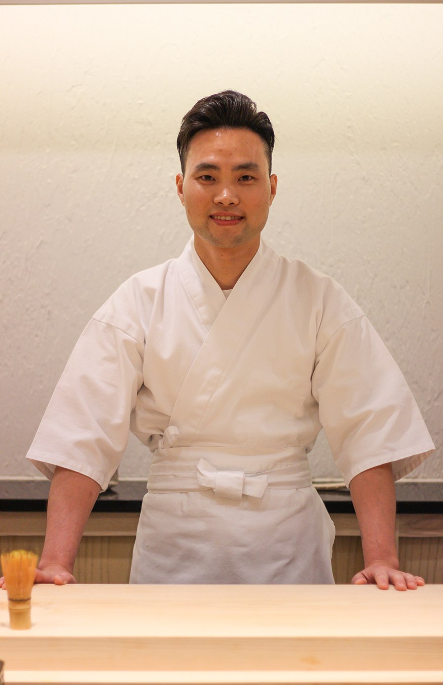 A man wearing chef's whites stands behind a light wooden counter, smiling at the camera.