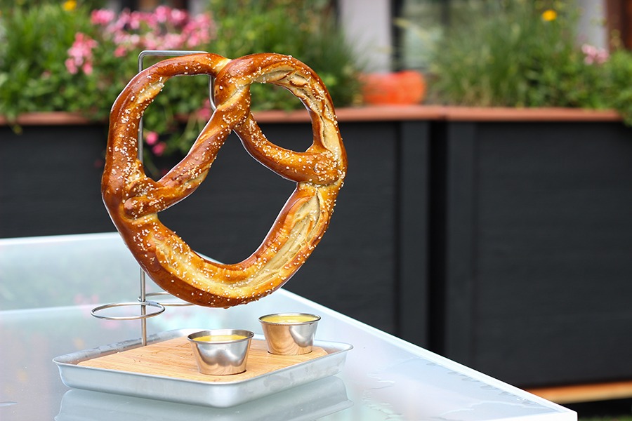 A very large pretzel hangs on a metal hook over two small metal cups of dipping sauces on a restaurant patio.