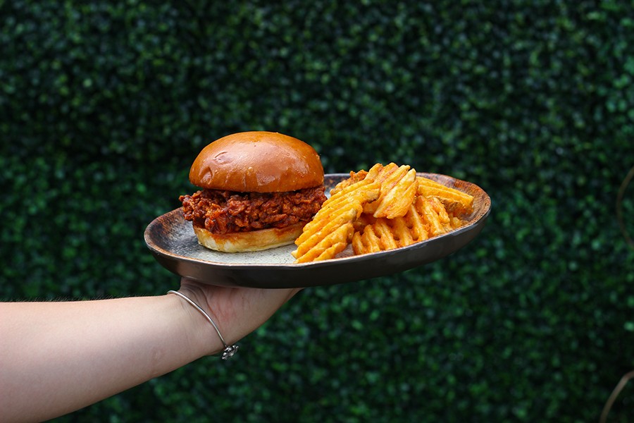 A hand holds up a plate in front of a greenery-covered wall. On the plate is a spicy, crispy fried chicken sandwich with a side of waffle fries.