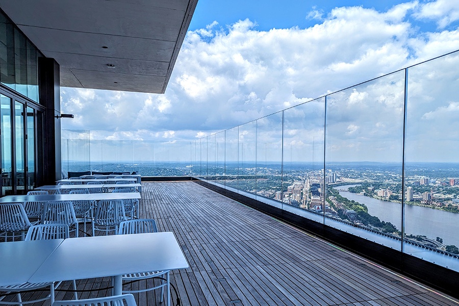 A roof deck has some chairs and tables, with glass barriers along the edge and skyline views all around.