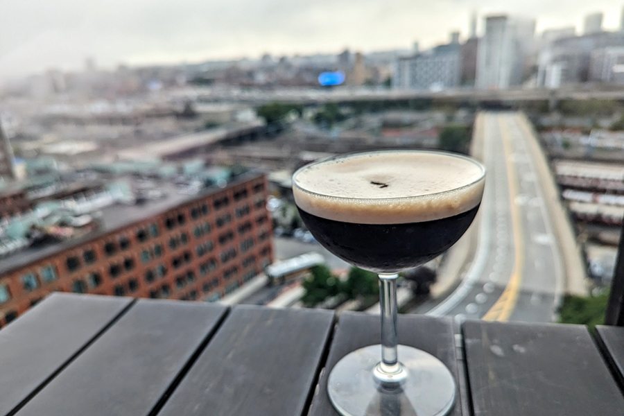 An espresso martini is on a wooden table on a roof deck, with a city skyline visible in the background.