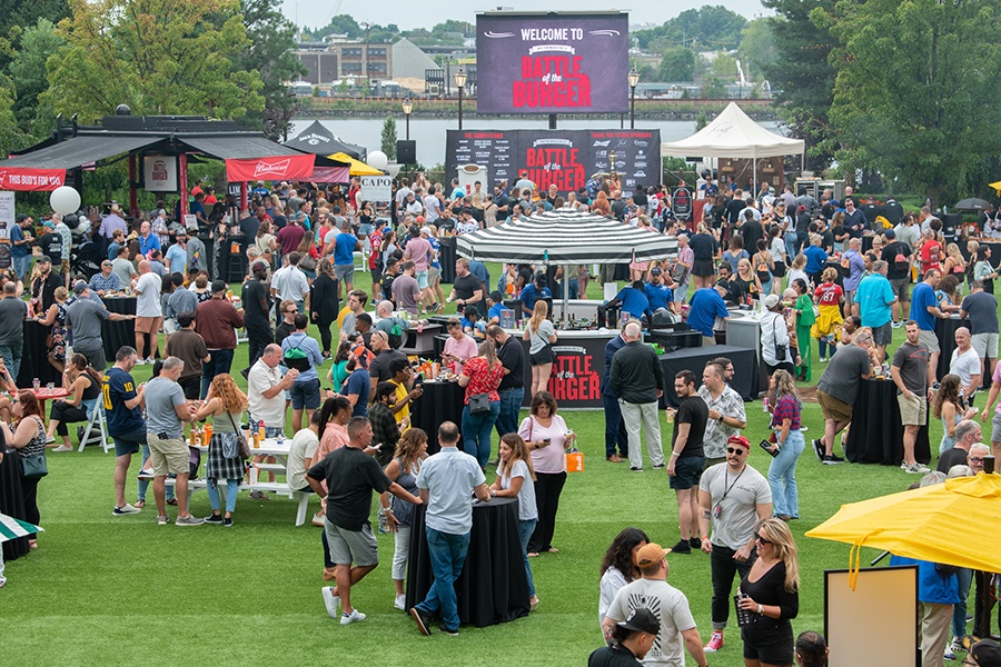 A casually dressed large crowd attends a daytime event on a large lawn, with Battle of the Burger branding visible.