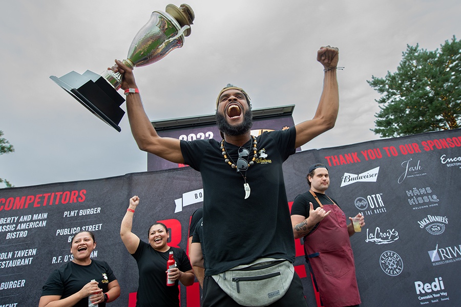 A man holds up a burger-shaped trophy triumphantly with several other people cheering in the background.