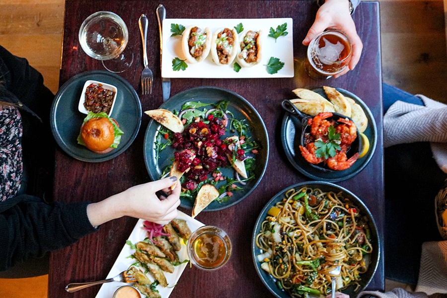 Overhead view of a table full of globally inspired small plates, with hands reaching in to take some.