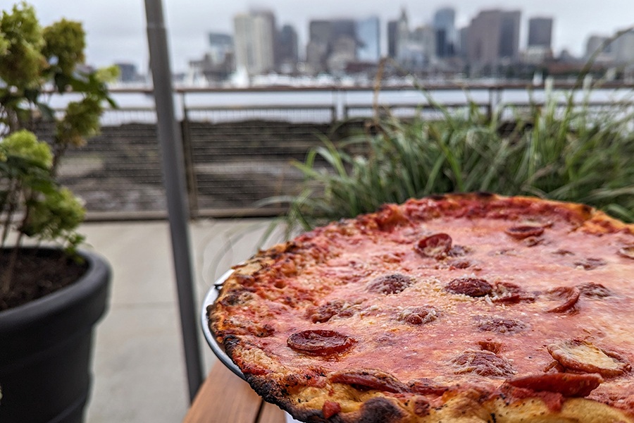 A thin-crust pepperoni pizza sits on a wooden table on a restaurant patio with water and a city skyline visible in the background.
