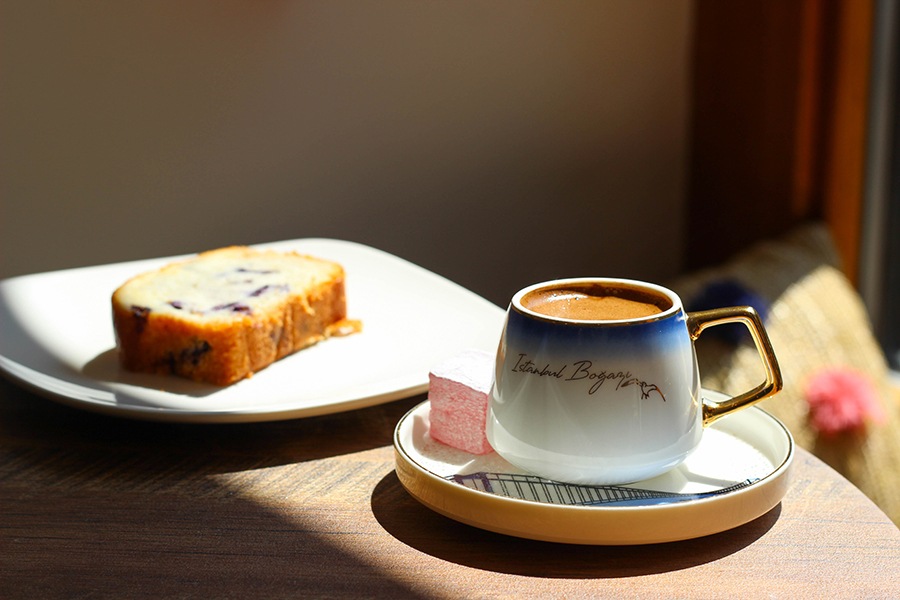 A blue, white, and gold mug is full of thick coffee and sits on a wooden table next to a slice of blueberry cake.