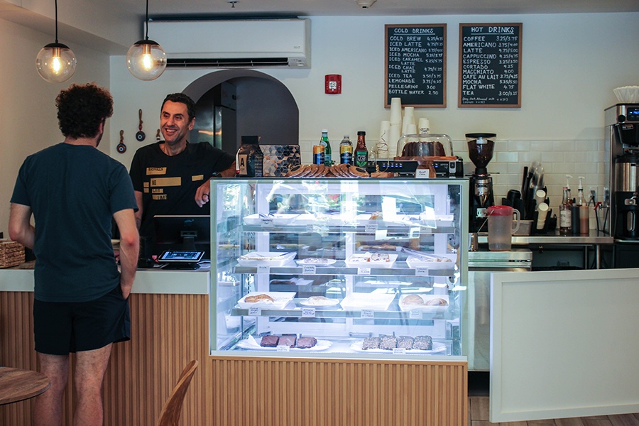 A man standing behind the register at a cafe smiles and takes a customer's order.