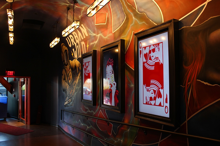 Framed Queen of Hearts-themed art is hung on the mural-covered wall of a tunnel-like entrance to an entertainment venue. The mural includes an astronaut DJ.