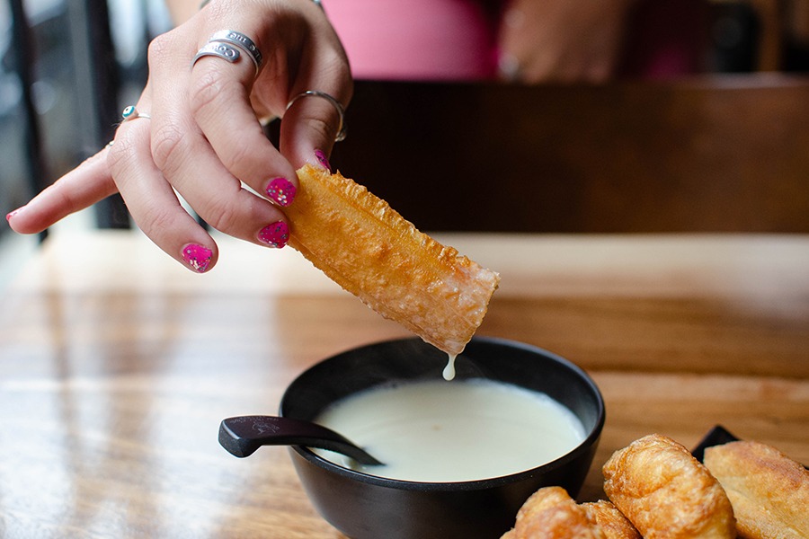 A hand dips a stick of fried dough into a bowl of a creamy white liquid.