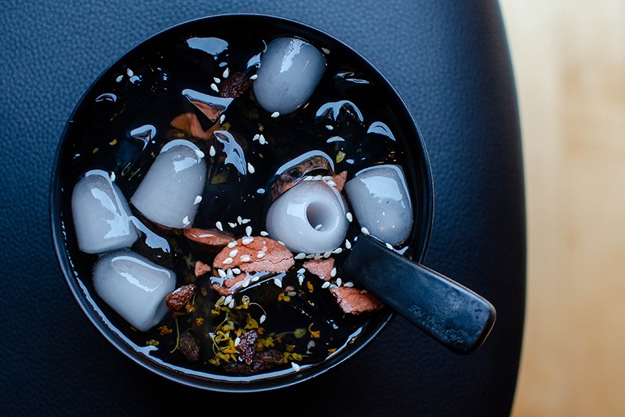 Overhead view of a bowl of a Chinese dessert, a clear jelly with ice cubes, sesame seeds, and dried floral bits.