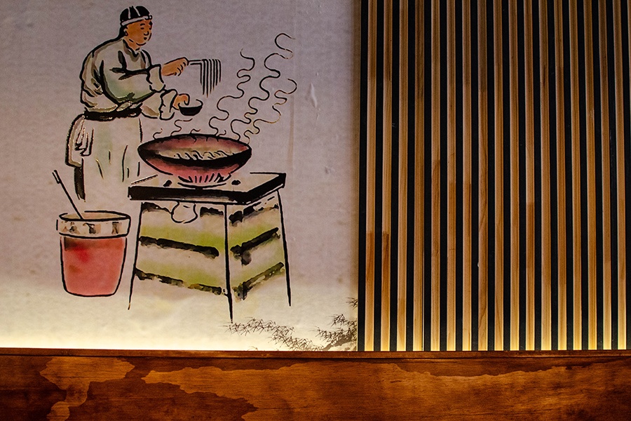 Art on the wall at a Chinese restaurant, featuring a man putting long noodles into a bowl from a large pot.