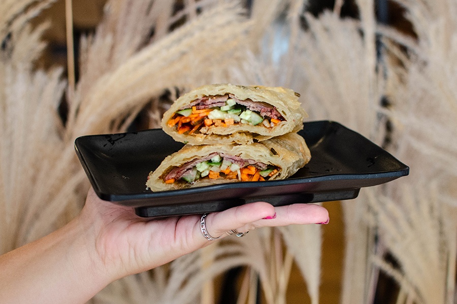 A hand holds up a black plate with two halves of a scallion pancake rollup stacked atop each other, stuffed with beef, cucumber, and carrot.