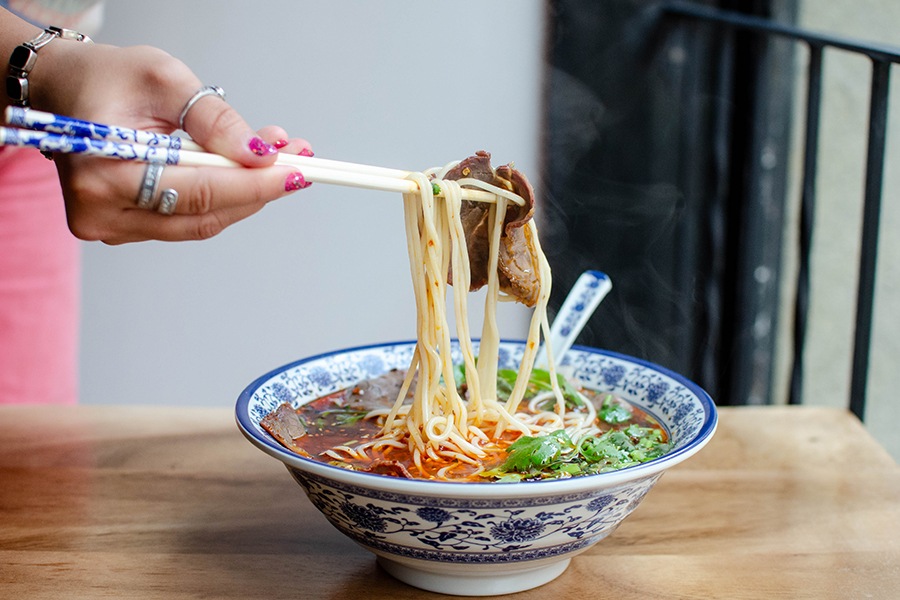 A hand uses chopsticks to pull noodles and a sliver of beef out of a steaming bowl of noodle soup, red with chili oil.