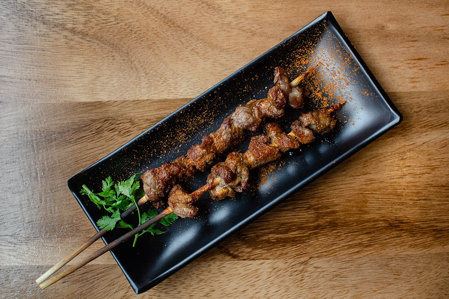 Overhead view of two skewers of grilled lamb chunks in a spicy red powder on a rectangular plate.