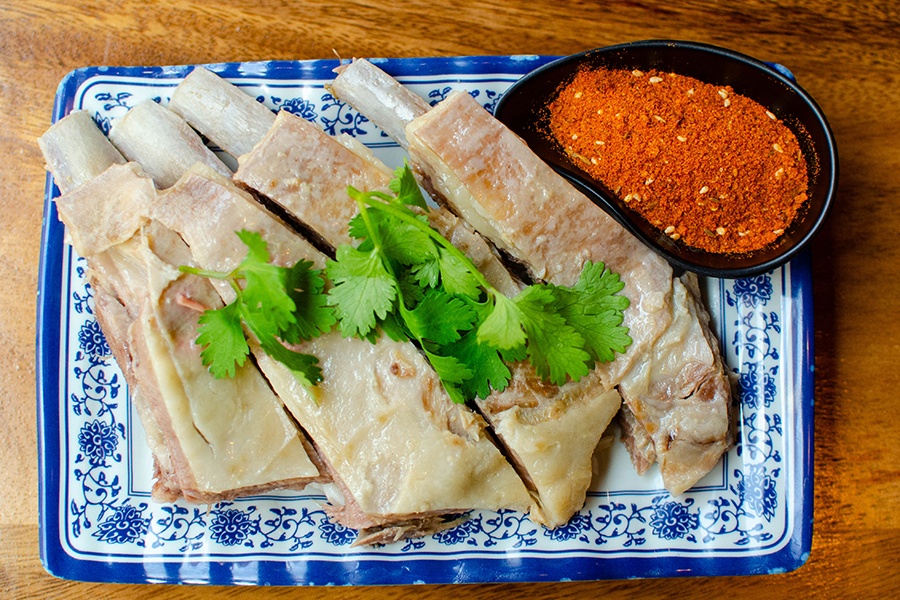 Four lamb chops sit on a blue floral rectangular plate with a small black bowl of a fiery red chili powder.