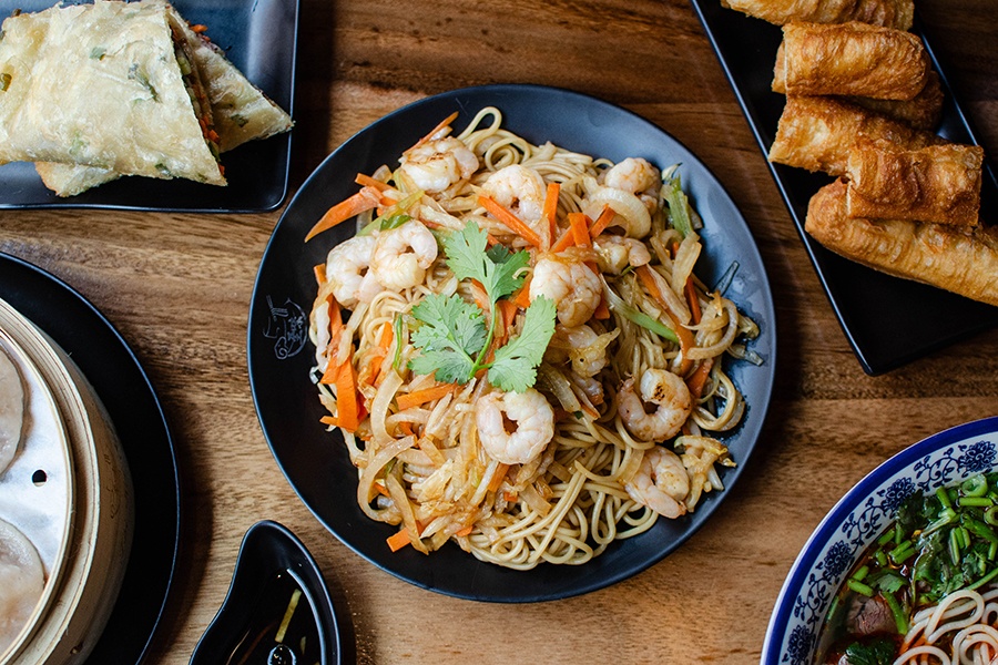 Overhead view of a noodle and shrimp dish surrounded by scallion pancake rollups, fried dough, a noodle soup, and more.