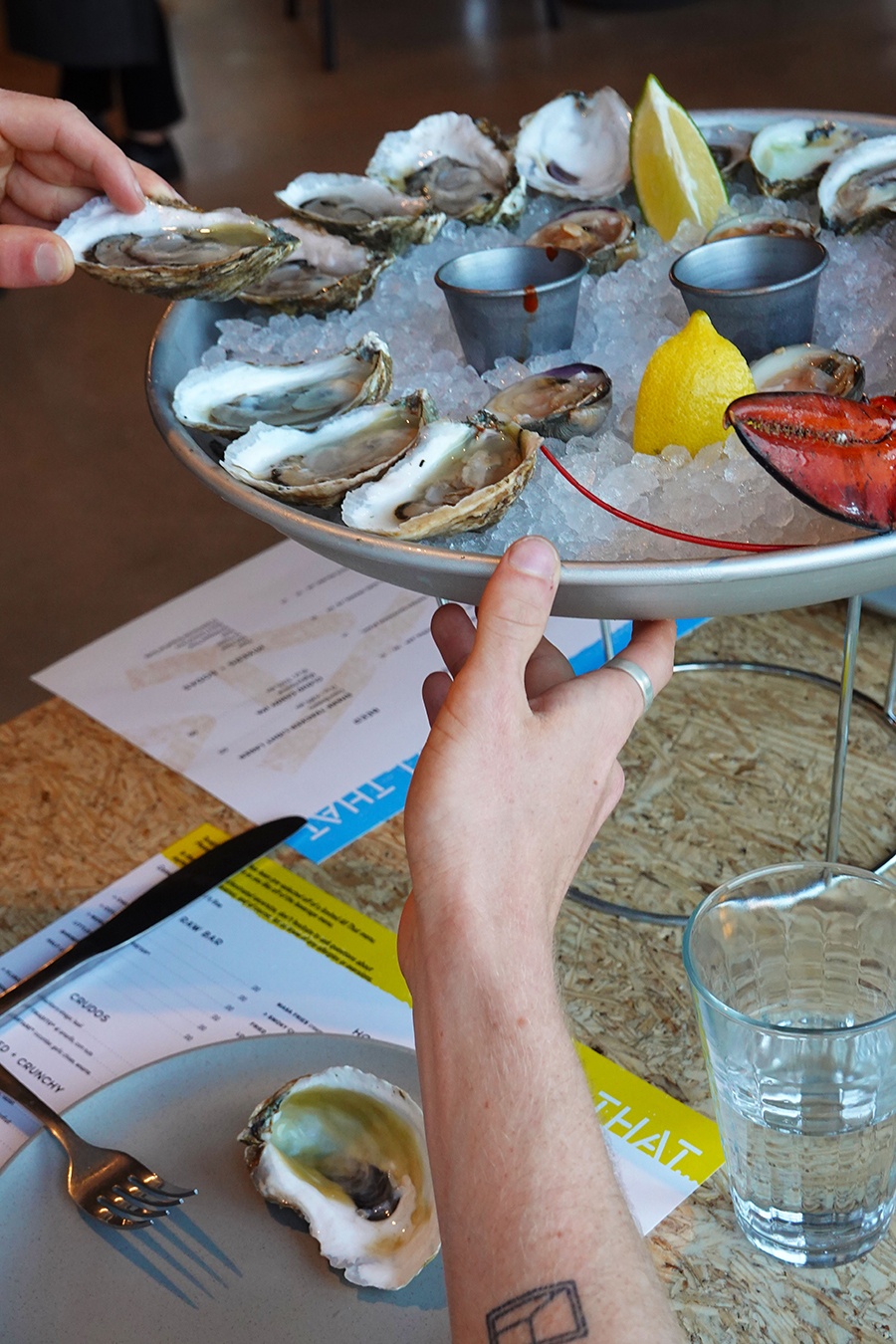 A hand grabs an oyster from a silver plate of ice and shellfish at a restaurant table.