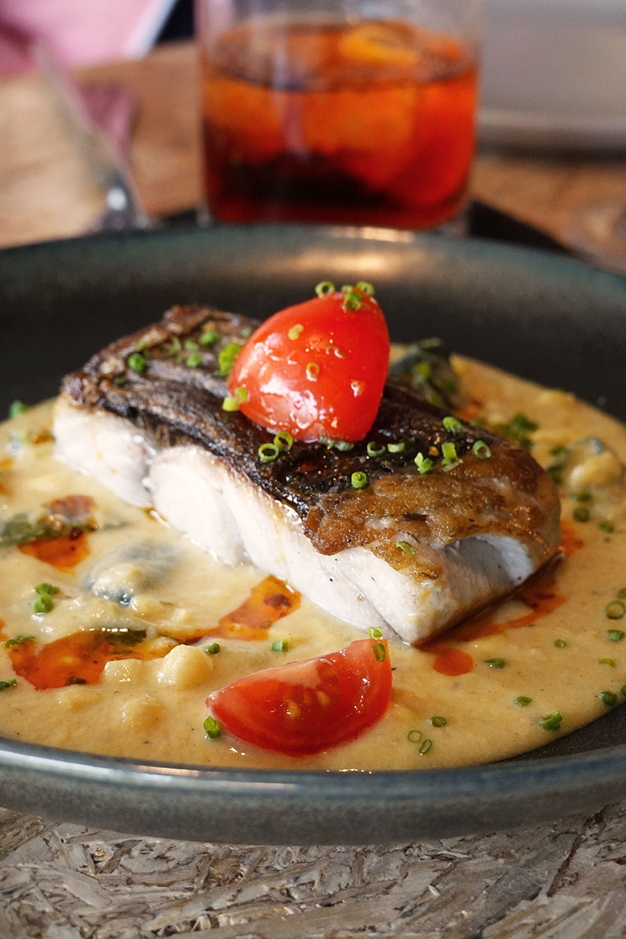 A thick filet of white fish with crispy skin is topped with chives and tomato and sits in a pool of pale yellow puree.