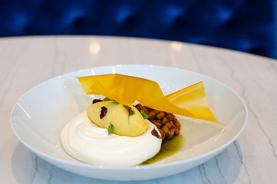 A dessert, plated in fine-dining style, features a quenelle of light orange sherbet on a thick cloud of whipped cream, topped with a curved sheet of dried mango.
