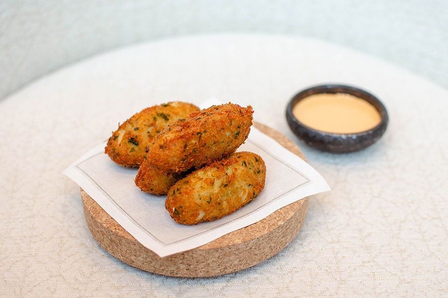 Four oblong, fried croquetas sit on a white napkin on a cork coaster with a little bowl of a pale orange dipping sauce to the side.