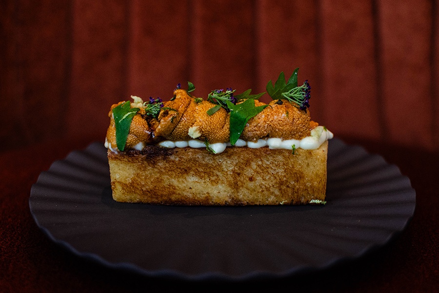 A rectangle of toast is topped with orange uni over a squiggle of white puree, garnished with green herbs and little purple flowers.