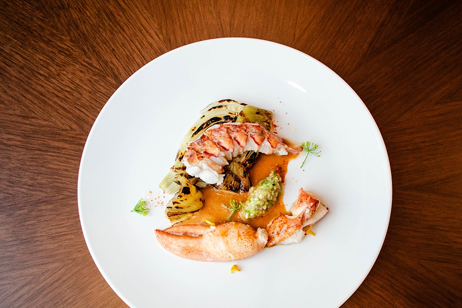 Overhead view of a lobster claw and tail on a plate with grilled pineapple and fennel and a thin brown sauce.