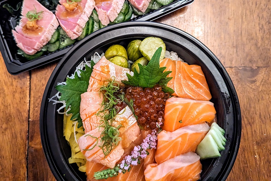 Overhead view of a salmon donburi bowl and seared tuna in black plastic takeover containers on a wooden table.