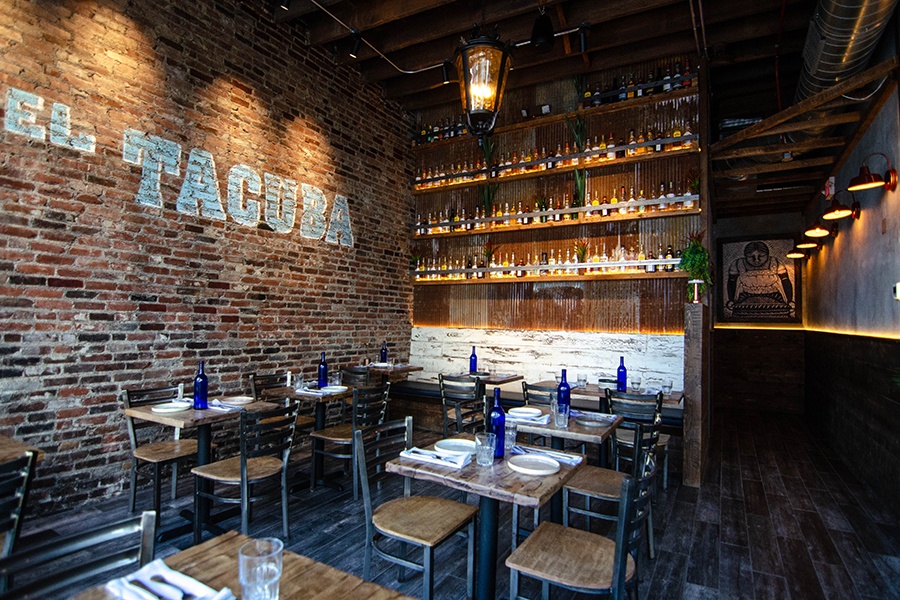 A restaurant interior features a wall of tequila bottles and the restaurant name, El Tacuba, painted on a brick wall.