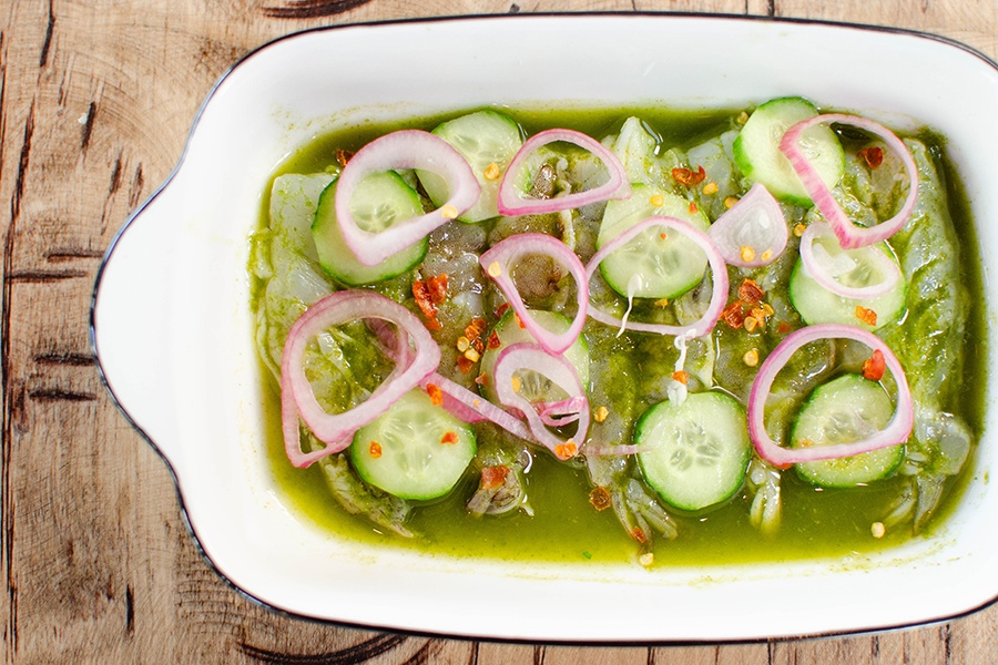Overhead view of a rectangular plate of shrimp ceviche in a thin green broth, topped with sliced red onions.