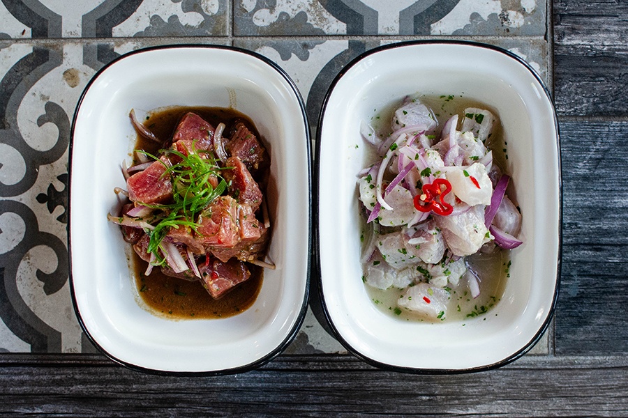 Overhead view of two raw fish dishes—one with tuna and one with white fish—on a weathered tile and wood surface.