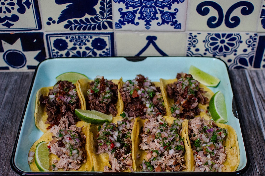 Eight tacos are lined up on an enamel tray in front of blue and white Talavera tiling.