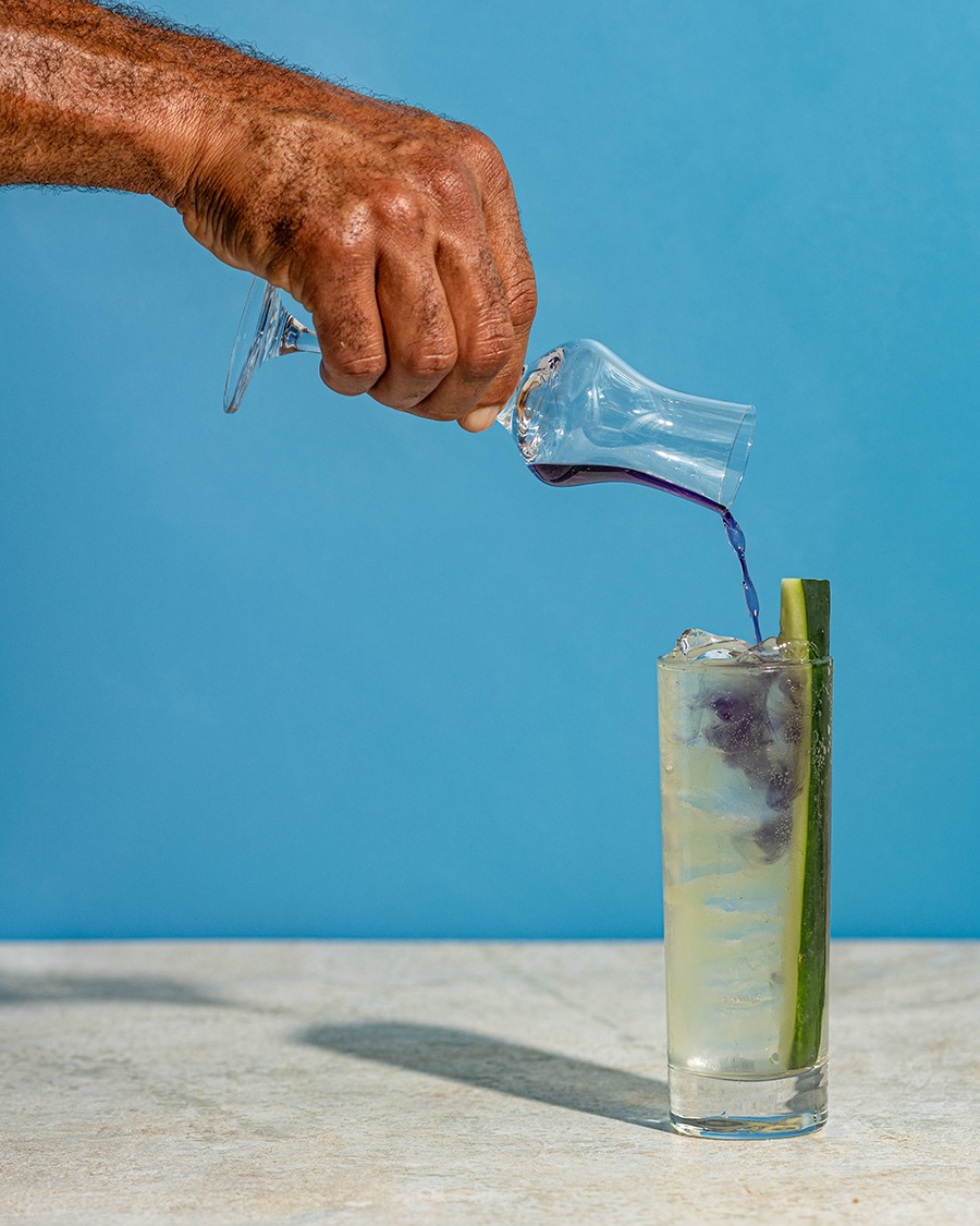 A hand pours purple liqueur into a clear cocktail garnished with a big piece of cucumber.