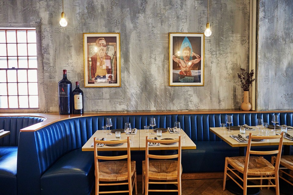 An interior shot of Krasi. There are gray and white plastered walls, two tables with four individual place settings, a royal blue seating banquette, a framed contemporary illustrations of Poseidon and Pythagoras.
