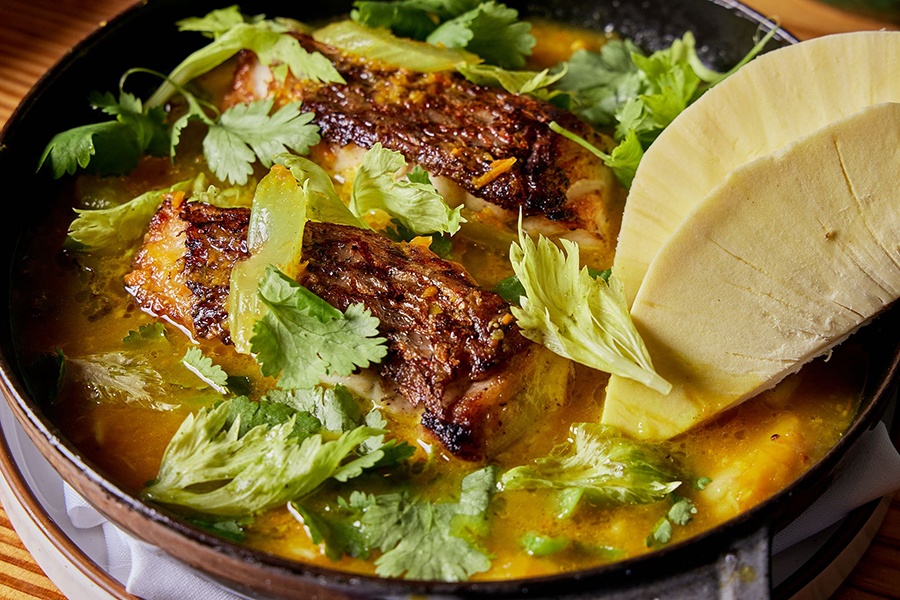 Closeup on a bowl of yellow soup with big pieces of fish with charred skin and lots of cilantro.