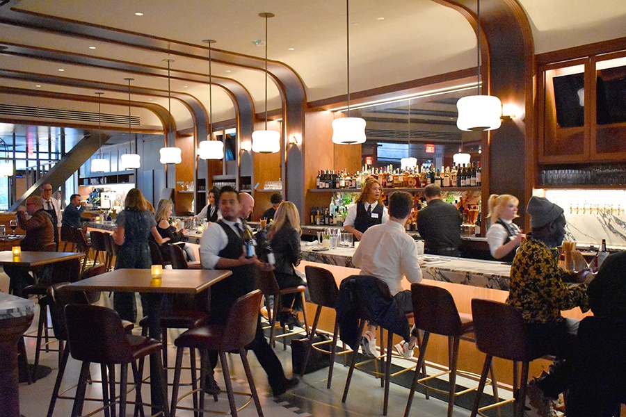 Patrons are gathered at a brasserie-style restaurant bar with lots of wood accents.