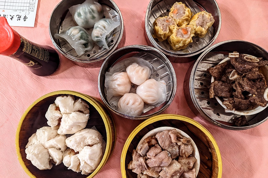 Overhead view of various dim sum dishes, including several types of dumplings, on a pale pink tablecloth.