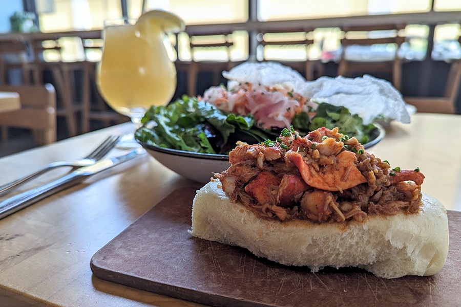 Lobster topped with chives sits in a fluffy bao-style bun on a wooden plank, with a salad and frozen drink in the background.