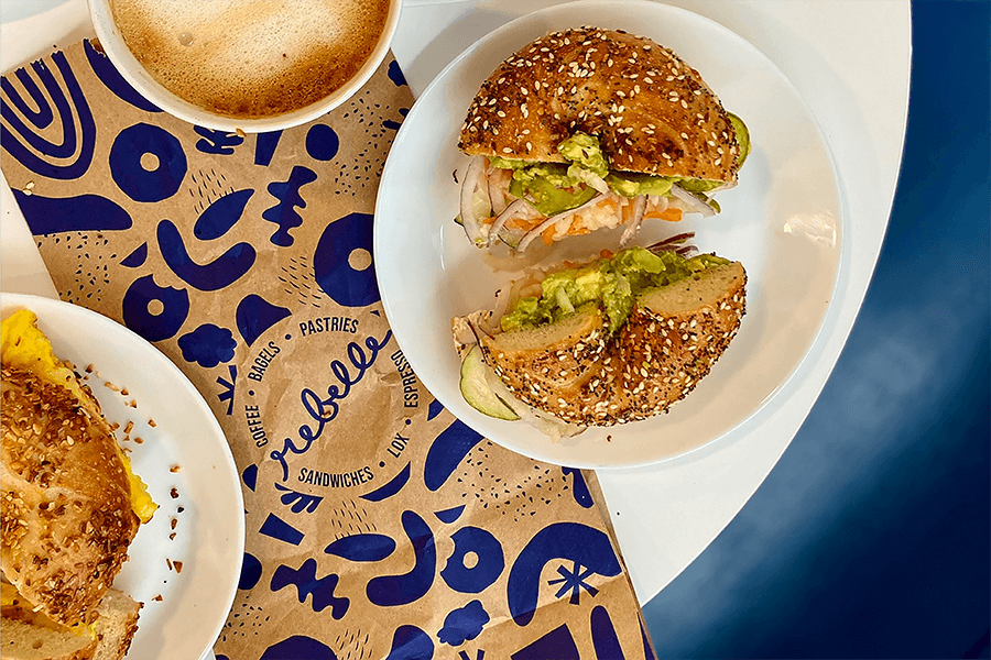 Overhead view of a couple bagel sandwiches and coffee with a paper bag that says Rebelle.