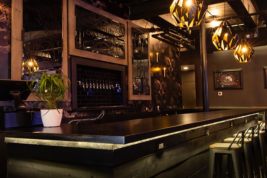 The bar inside a taproom features black walls and surfaces, angular goth light fixtures, and skull-themed art.