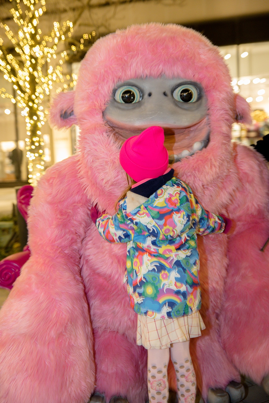 A child in a colorful unicorn winter jacket hugs a giant pink yeti.