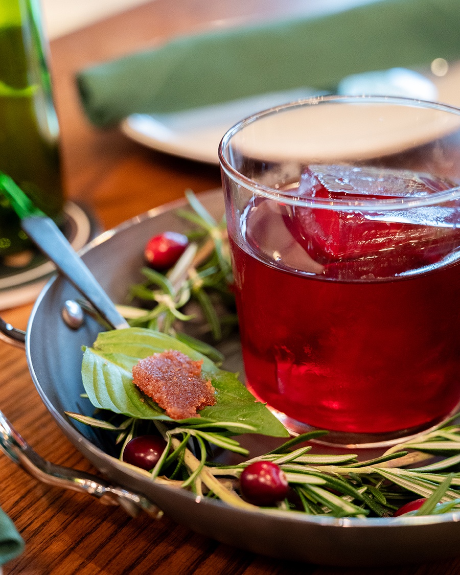 A red cocktail with a big ice cube sits on a plate of cranberries and rosemary.