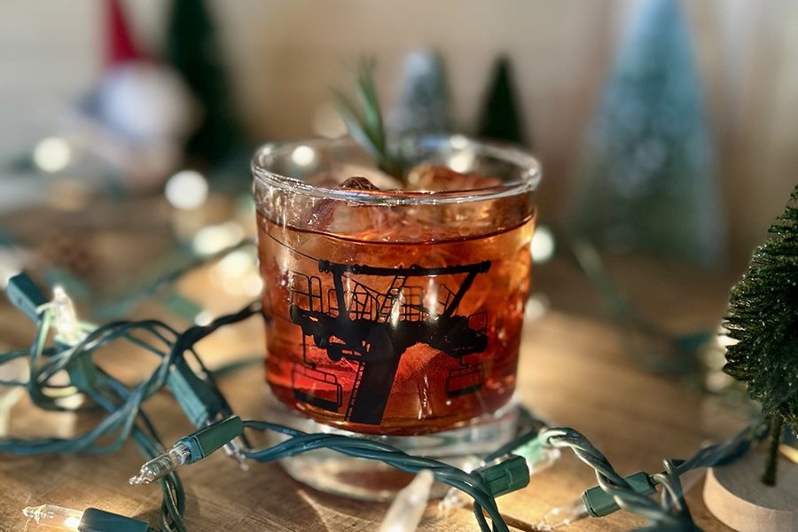 A brown cocktail is in a short glass decorated with the silhouette of a ski lift. Christmas lights and fake trees surround the glass.