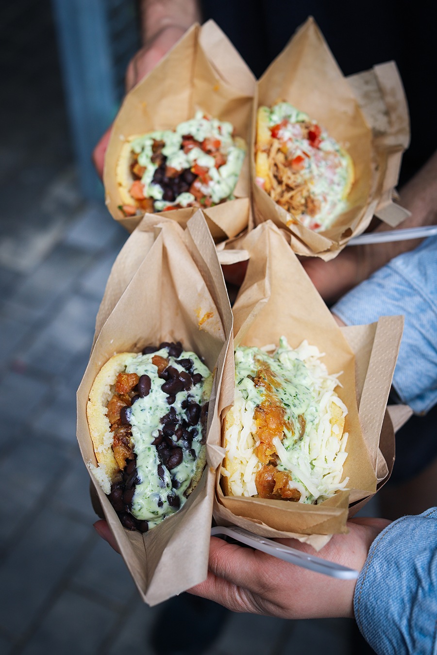 Two pairs of hands hold two arepas each, stuffed full of various fillings.