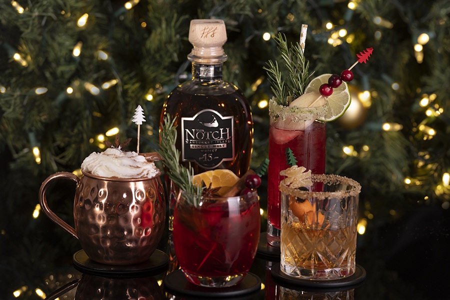 A few cocktails with Christmas-y garnishes, like rosemary and cranberries, sit in front of a Christmas tree.