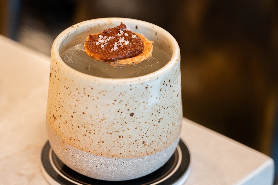 A light-colored cocktail in a speckled earthenware cup has one large ice cube in it and a mole-topped cracker as garnish.