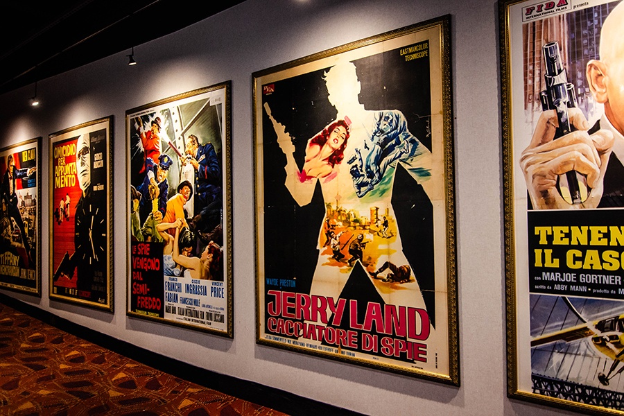 Old spy film posters in Italian line the walls of a movie theatre hallway.