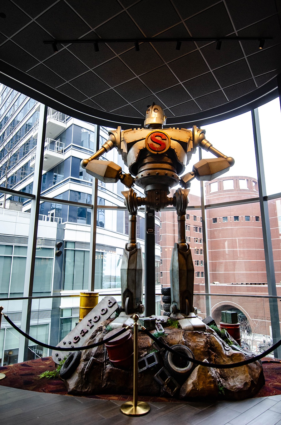 A tall metal sculpture of a giant stands on a pile of debris in a high-ceilinged building lobby with a city skyline visible through the windows behind.