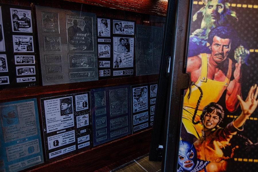 Old film poster printing plates are displayed on a wall near a colorful sandwich board advertising newer films at a movie theatre.