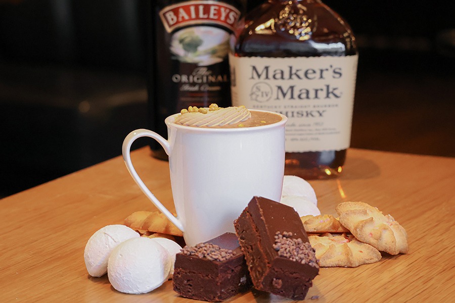 A white mug is full of hot chocolate and surrounded by sweet treats. Bottles of Baileys and Maker's Mark are visible in the background.