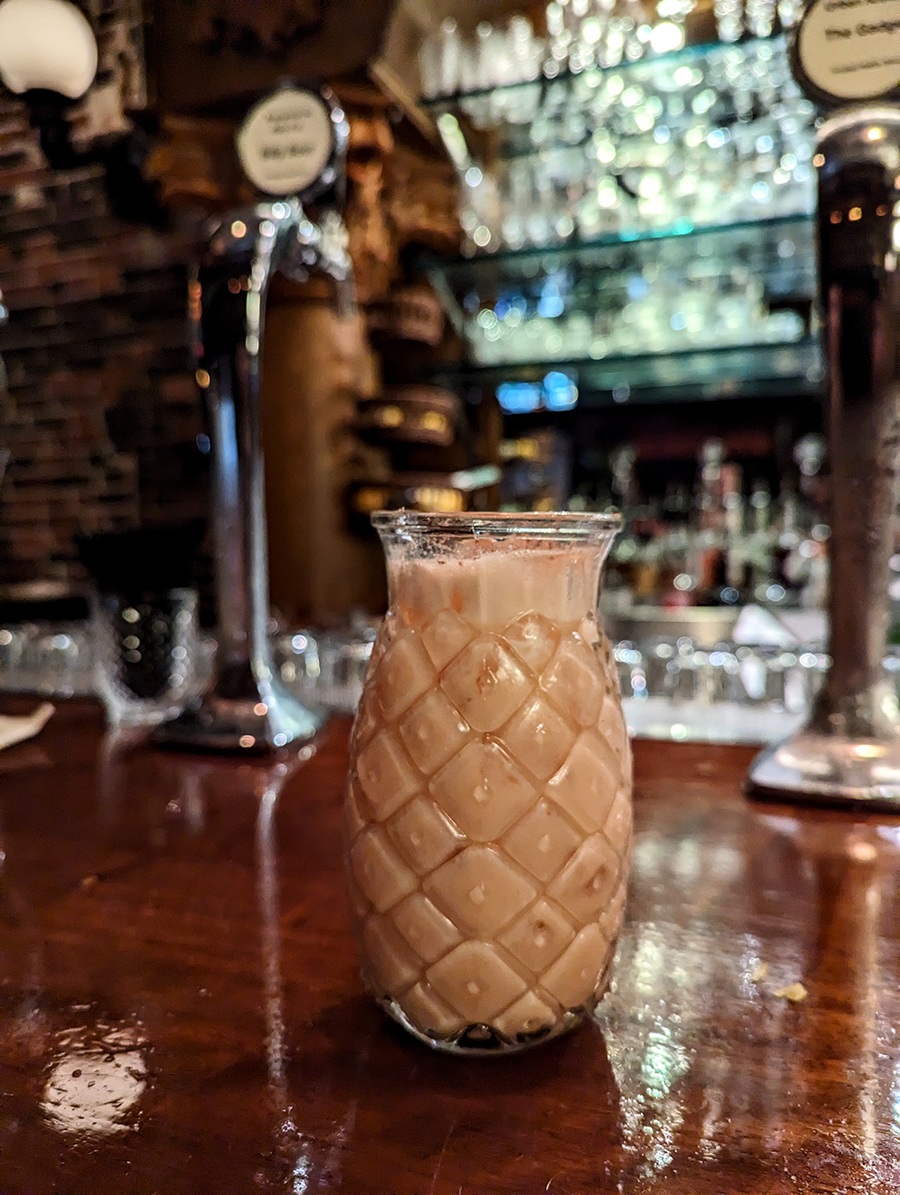 A foamy pink cocktail is served in pineapple-shaped glassware on a glossy wooden bar.